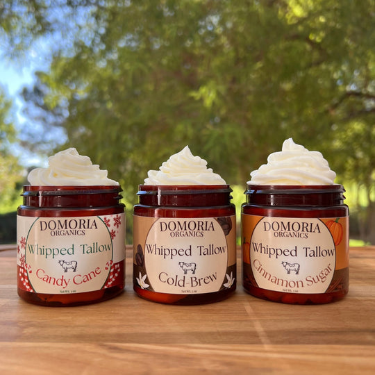 Three jars of Domoria Organics whipped tallow products on a wooden surface with a natural background.